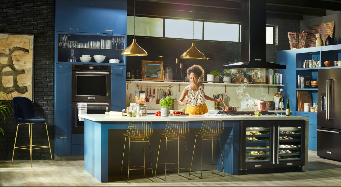 Person cooking in a kitchen with deep blue cabinets and black stainless steel KitchenAid® appliances Person cooking in a kitchen with deep blue cabinets and black stainless steel KitchenAid® appliances