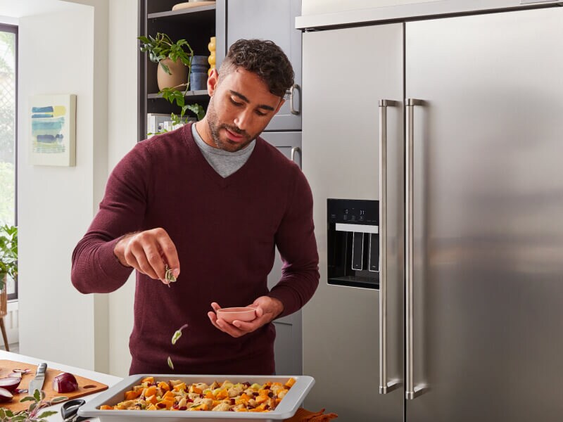Person seasoning food in front of a stainless steel side-by-side refrigerator Person seasoning food in front of a stainless steel side-by-side refrigerator