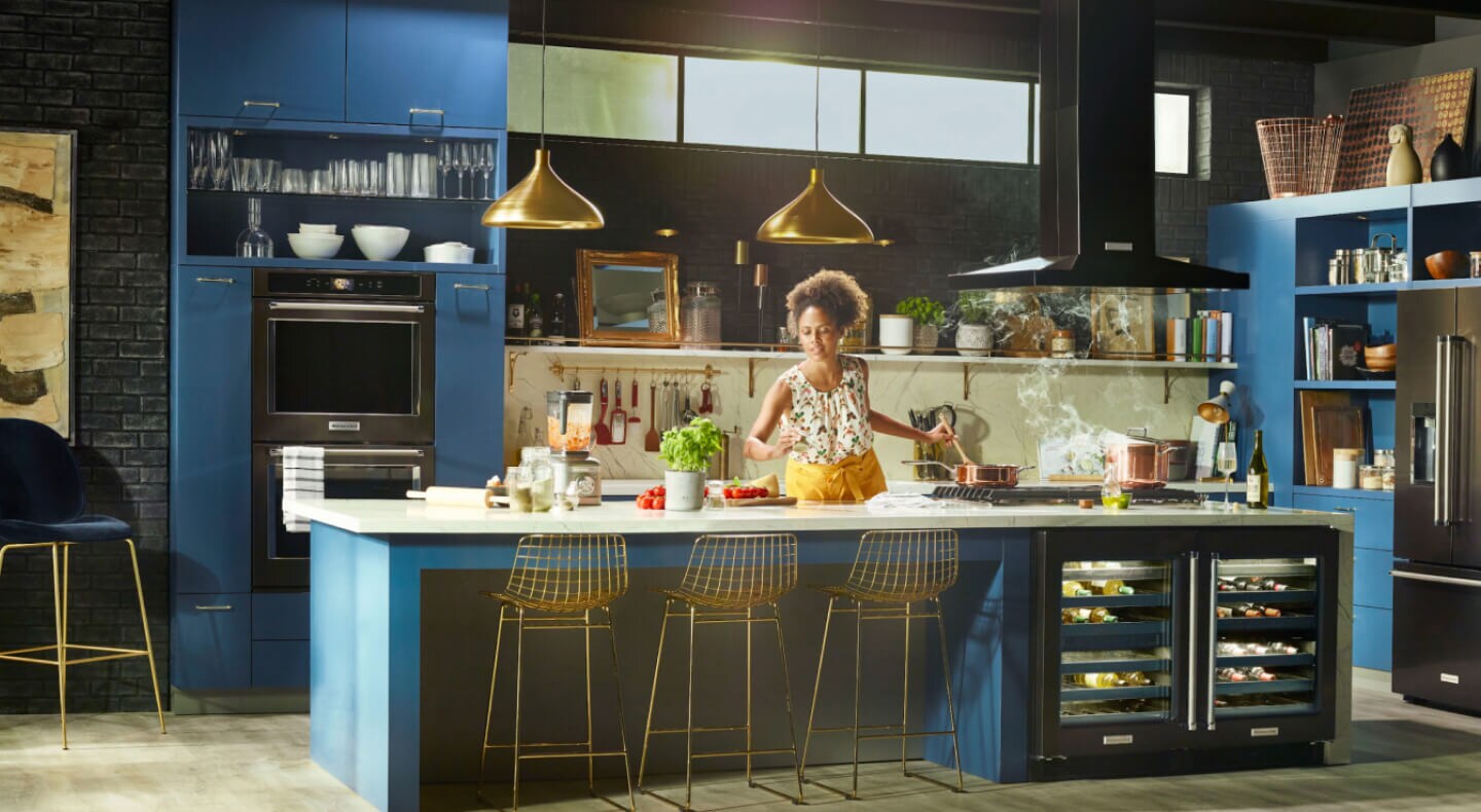 Person cooking in a kitchen with deep blue cabinets and black stainless steel KitchenAid® appliances Person cooking in a kitchen with deep blue cabinets and black stainless steel KitchenAid® appliances