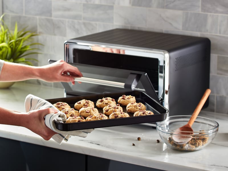 Person placing a tray of cookies into a KitchenAid® countertop oven Person placing a tray of cookies into a KitchenAid® countertop oven