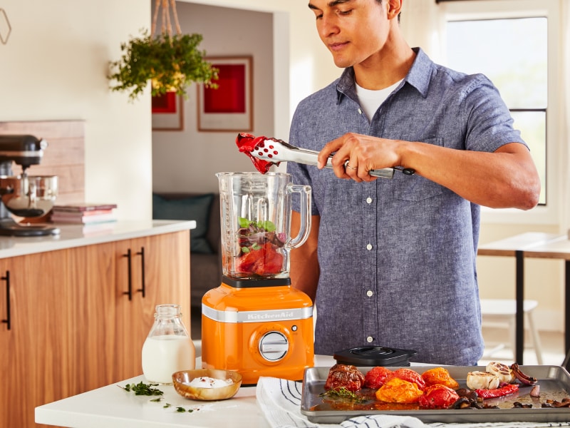A man putting roasted vegetables into a KitchenAid® blender in a modern kitchen. A man putting roasted vegetables into a KitchenAid® blender in a modern kitchen.