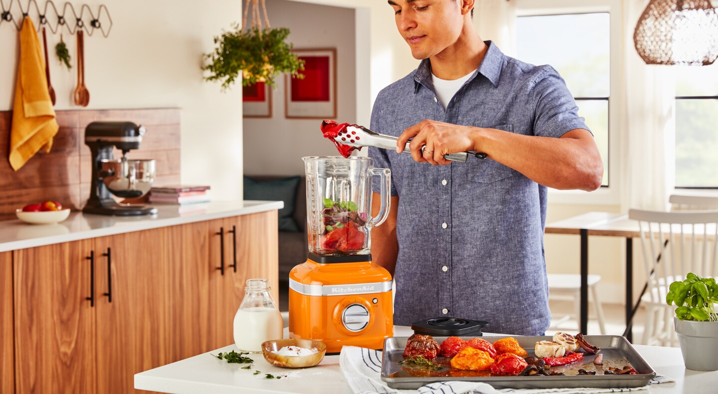 A man putting roasted vegetables into a KitchenAid® blender in a modern kitchen. A man putting roasted vegetables into a KitchenAid® blender in a modern kitchen.