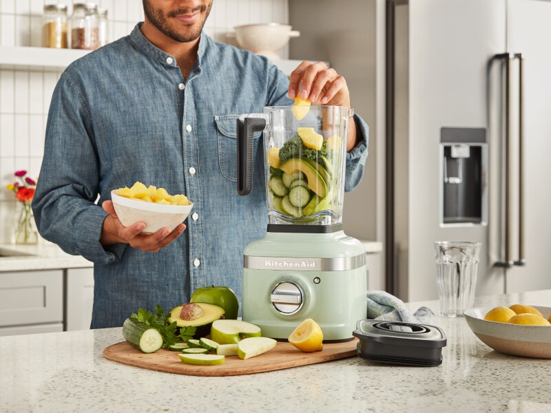 A man adding vegetables to a KitchenAid® blender in a modern kitchen. A man adding vegetables to a KitchenAid® blender in a modern kitchen.