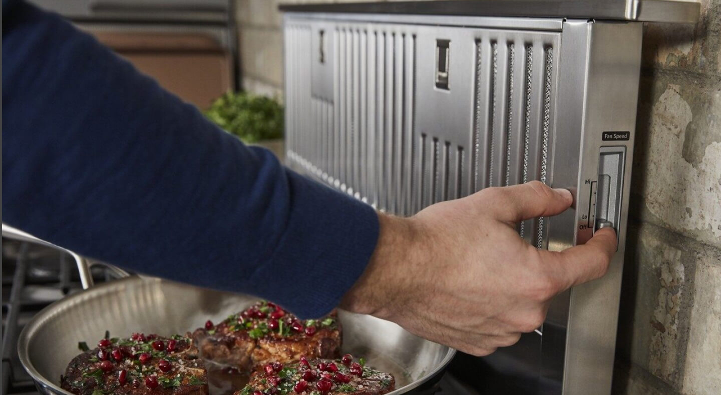 Man turning on a downdraft vent while meat cooks on the stove Man turning on a downdraft vent while meat cooks on the stove