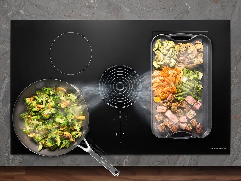 Overhead view of an induction cooktop with multiple dishes cooking and steam being pulled down the downdraft vent