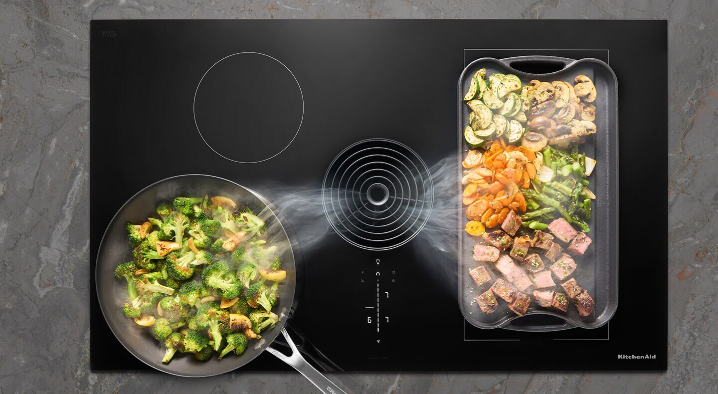 Overhead view of an induction cooktop with multiple dishes cooking and steam being pulled down the downdraft vent