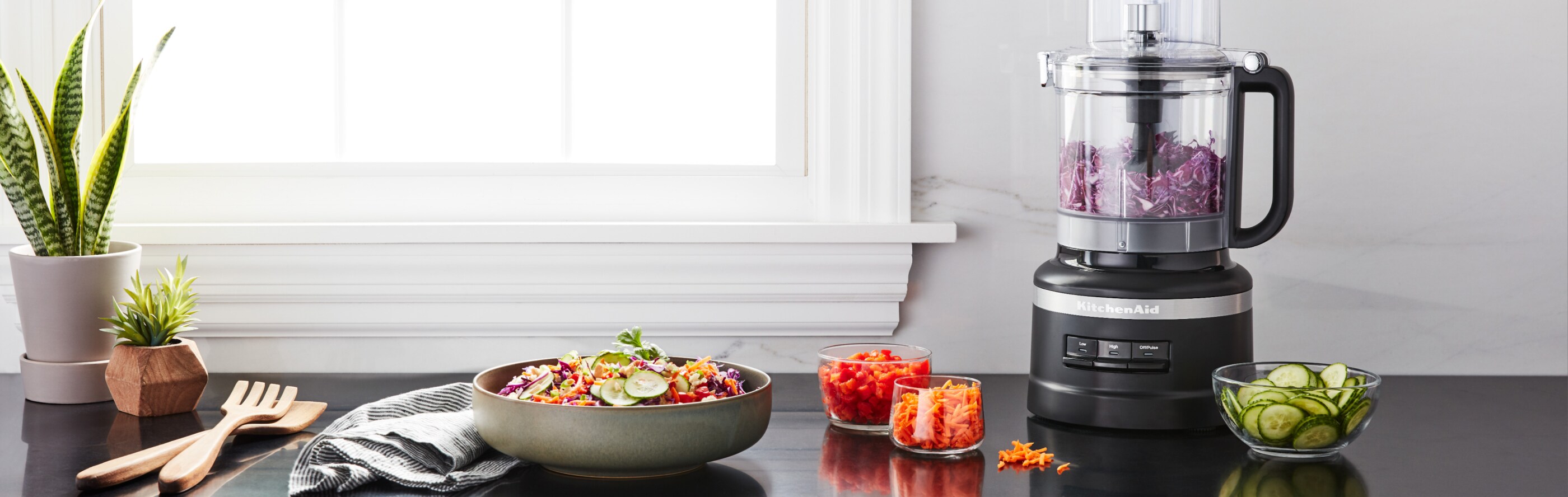Black KitchenAid® food processor full of shredded red onion next to prepared veggies on the countertop