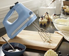 A KitchenAid 5-Speed Hand Mixer on a cutting board surrounded by frosting, jam and cupcakes.