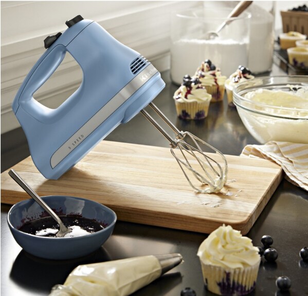 A KitchenAid 5-Speed Hand Mixer on a cutting board surrounded by frosting, jam and cupcakes.