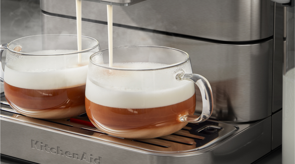 A pair of glass espresso cups filled with espresso, with steamed milk pouring into the glasses from the machine's dual spouts.