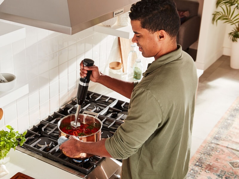 A person using a KitchenAid® hand blender to blend food in a saucepan on a gas cooktop