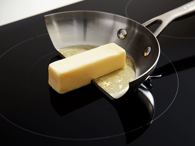 Half of a pan with a stick of butter half on the cooktop and half off, only melting in the pan, showing the relative coolness of an induction stovetop without the cookware