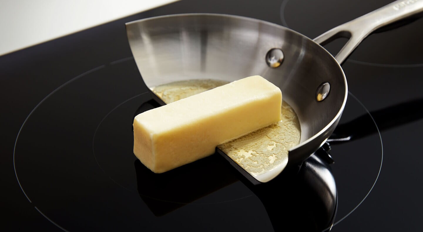 Half of a pan with a stick of butter half on the cooktop and half off, only melting in the pan, showing the relative coolness of an induction stovetop without the cookware
