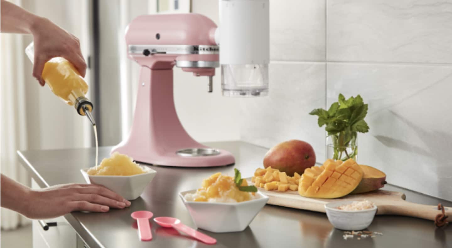 Person pouring syrup on a bowl of shaved ice surrounded by KitchenAid® stand mixer and chopped mangoes Person pouring syrup on a bowl of shaved ice surrounded by KitchenAid® stand mixer and chopped mangoes