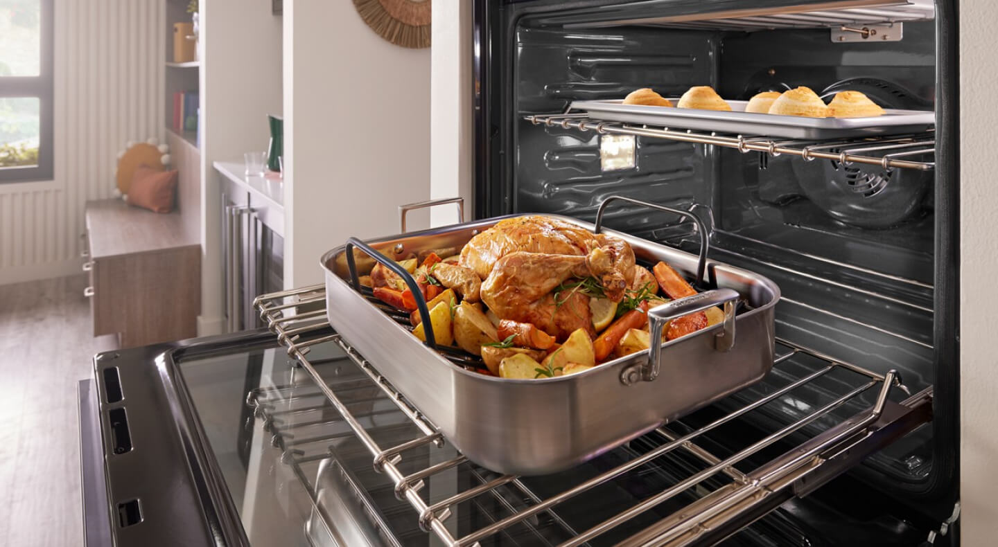 A pulled back image of a kitchen with an open oven in it with a chicken on the bottom rack and bread rolls on the top rack