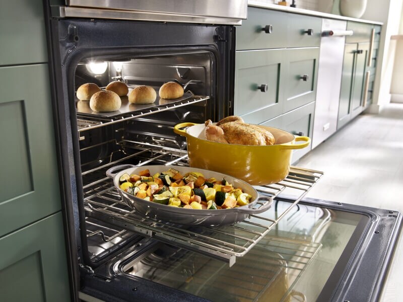 A close up of an open oven in a kitchen with a chicken, vegetables and bread rolls all cooking inside