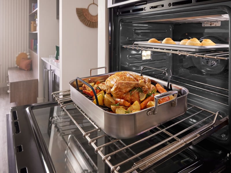 A pulled back image of a kitchen with an open oven in it with a chicken on the bottom rack and bread rolls on the top rack