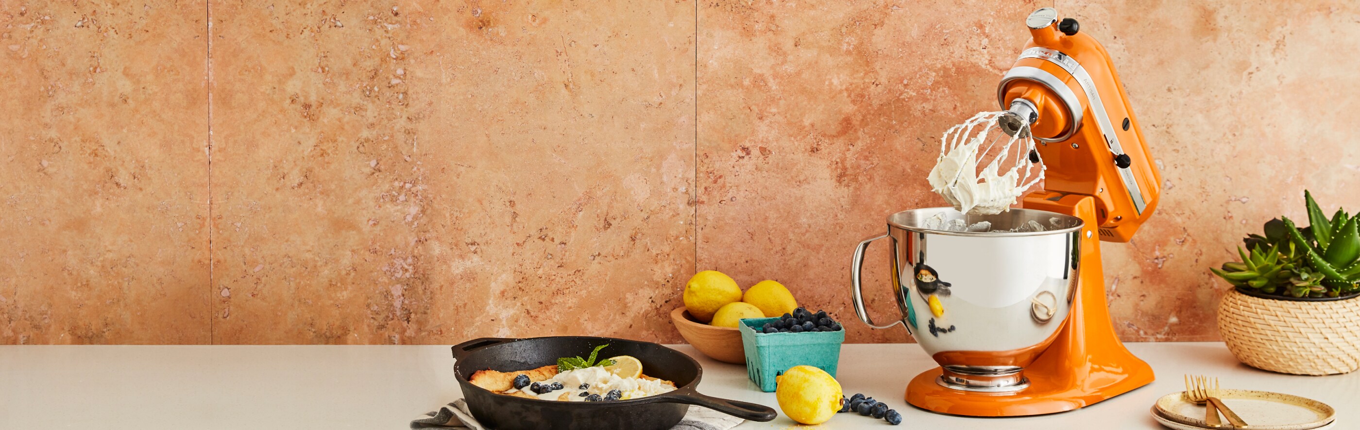 An orange KitchenAid® stand mixer, fruit and a cast iron pan.