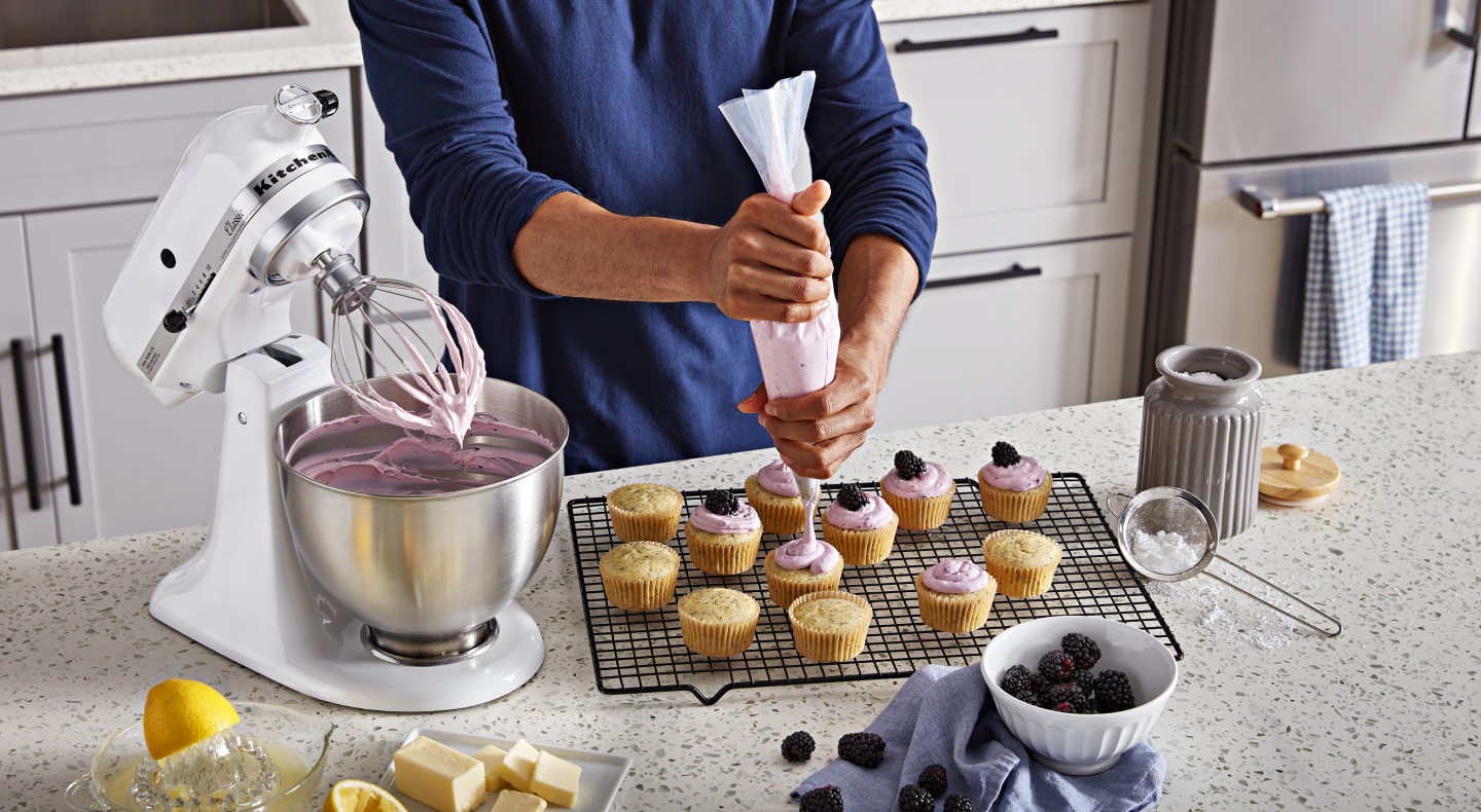 Person piping frosting onto cupcakes next to white stand mixer full of frosting Person piping frosting onto cupcakes next to white stand mixer full of frosting