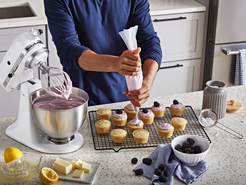 Person piping frosting onto cupcakes next to white stand mixer full of frosting Person piping frosting onto cupcakes next to white stand mixer full of frosting