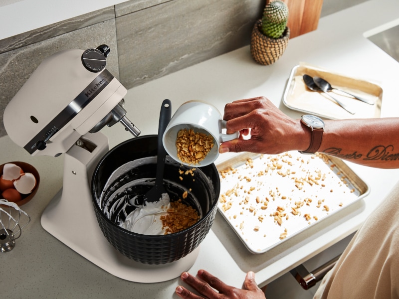 Person adding ingredients to black stand mixer bowl attached to white stand mixer