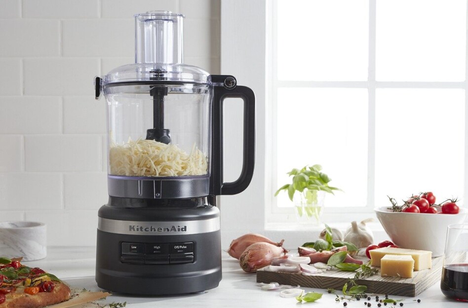 Black KitchenAid brand food processor with shredded cheese next to cutting board and flatbread Black KitchenAid brand food processor with shredded cheese next to cutting board and flatbread
