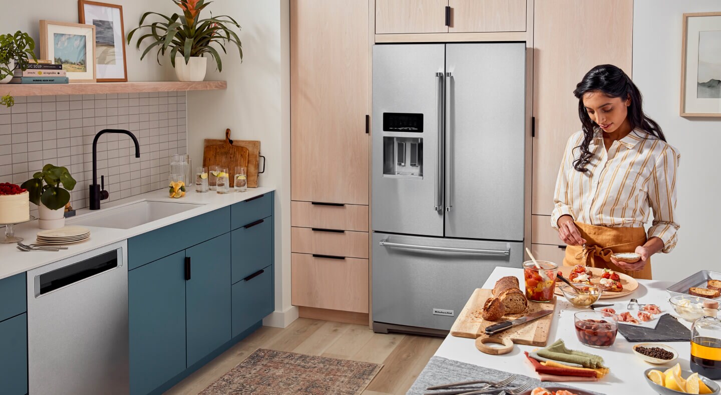 A person prepping food at an island with a bread and cheese spread in a kitchen with blue cabinets and a French door refrigerator A person prepping food at an island with a bread and cheese spread in a kitchen with blue cabinets and a French door refrigerator