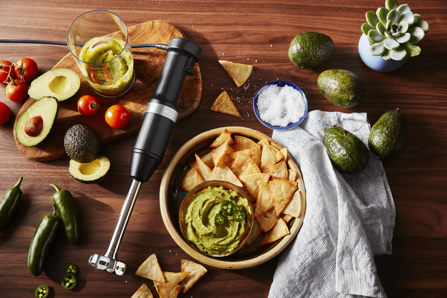 Black KitchenAid® immersion blender on wood surface with guacamole ingredients and blended guacamole with chips Black KitchenAid® immersion blender on wood surface with guacamole ingredients and blended guacamole with chips