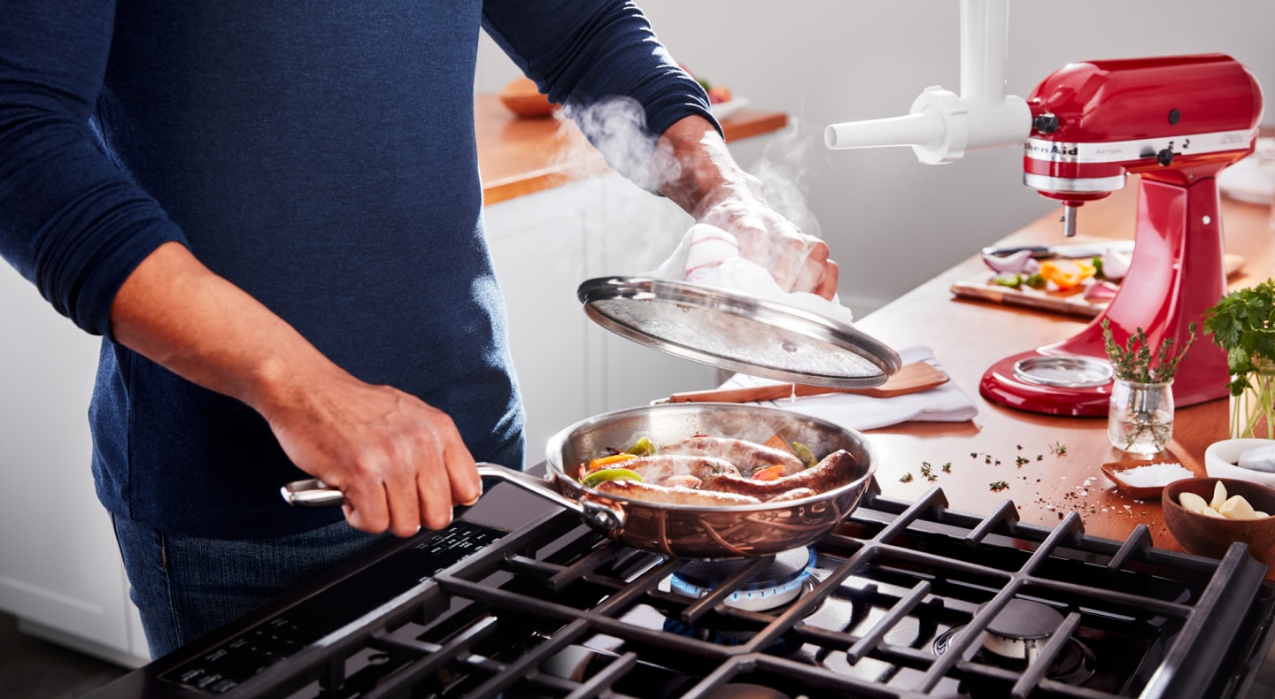 Person cooking sausages over a gas range with herbs, veggies and KitchenAid® stand mixer on the countertop Person cooking sausages over a gas range with herbs, veggies and KitchenAid® stand mixer on the countertop