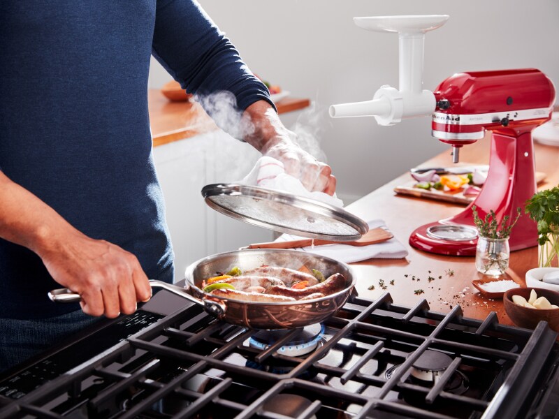 Person cooking sausages over a gas range with herbs, veggies and KitchenAid® stand mixer on the countertop Person cooking sausages over a gas range with herbs, veggies and KitchenAid® stand mixer on the countertop