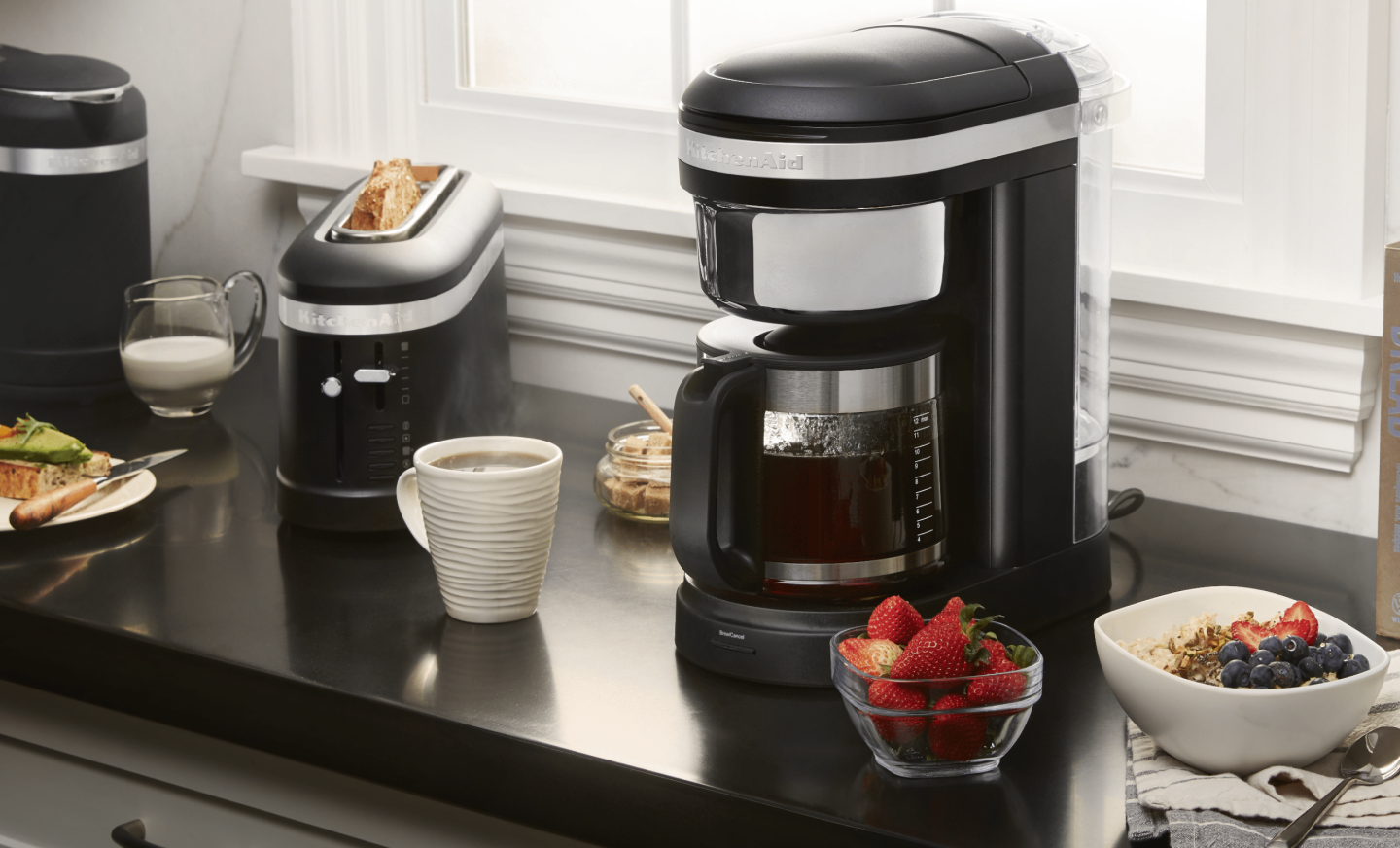 KitchenAid® coffee maker on countertop with coffee drink and breakfast foods KitchenAid® coffee maker on countertop with coffee drink and breakfast foods