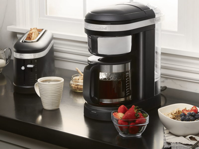 KitchenAid® coffee maker on countertop with coffee drink and breakfast foods KitchenAid® coffee maker on countertop with coffee drink and breakfast foods