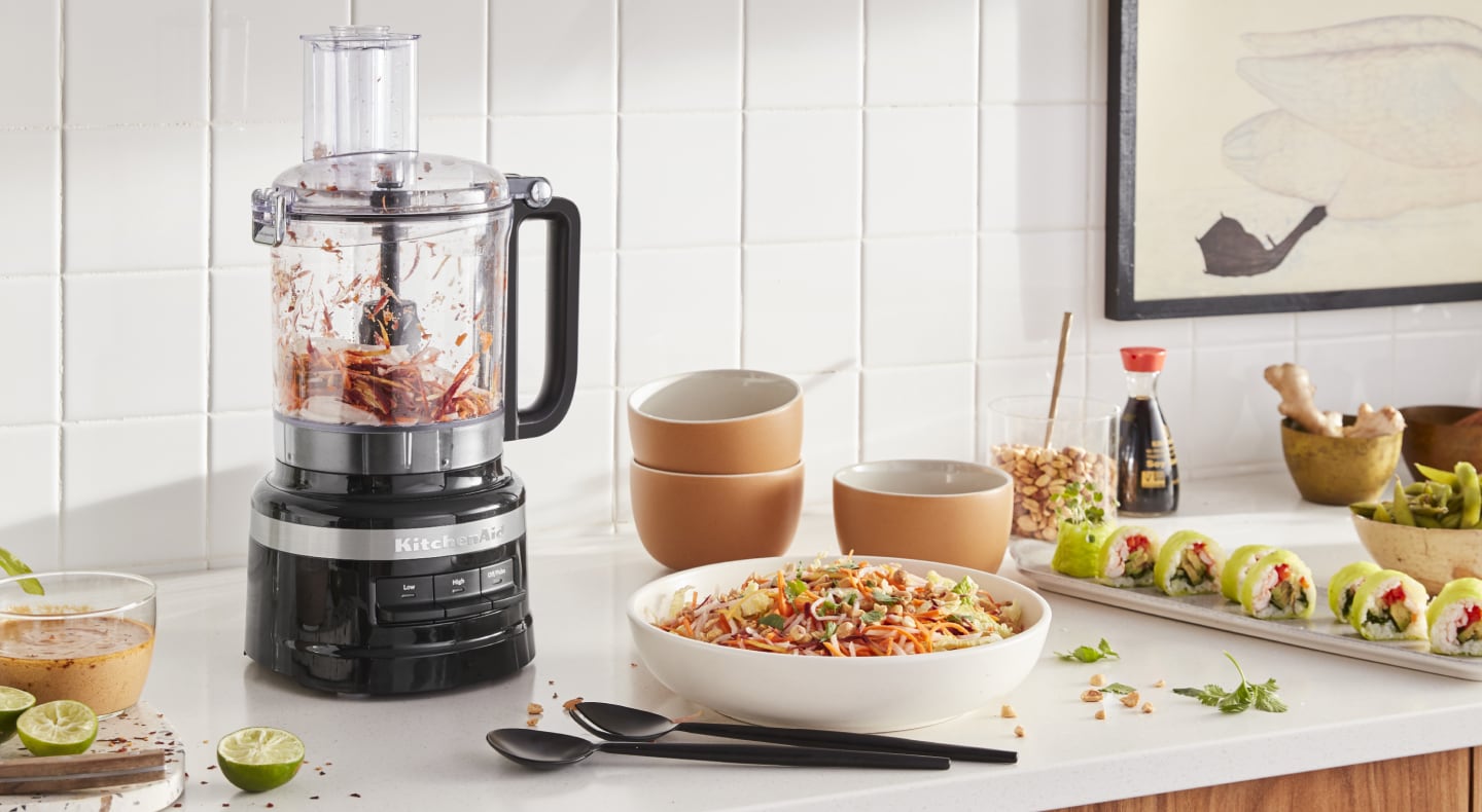 KitchenAid® food processor with chopped vegetables next to a chopped salad and freshly made salad dressing in a modern kitchen. KitchenAid® food processor with chopped vegetables next to a chopped salad and freshly made salad dressing in a modern kitchen.