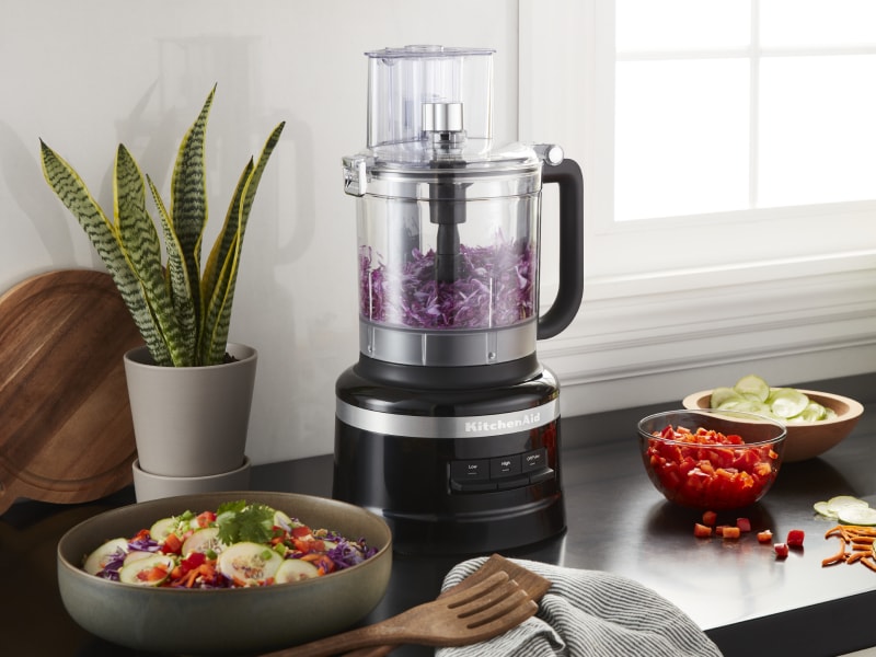 KitchenAid® food processor with purple cabbage next to a chopped salad and the ingredients in a modern kitchen. KitchenAid® food processor with purple cabbage next to a chopped salad and the ingredients in a modern kitchen.