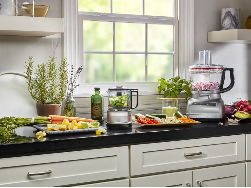 KitchenAid® food processor and food chopper with chopped vegetables next to fresh vegetables in a modern kitchen. KitchenAid® food processor and food chopper with chopped vegetables next to fresh vegetables in a modern kitchen.