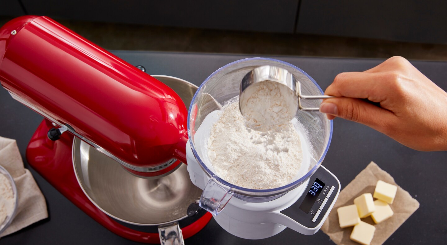 Person pouring flour from a measuring cup into the KitchenAid® Sifter + Scale Attachment
