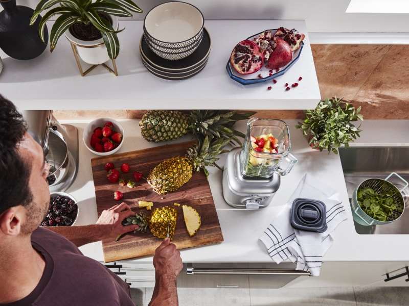 Person cutting pineapple next to KitchenAid® blender with fruit in it Person cutting pineapple next to KitchenAid® blender with fruit in it
