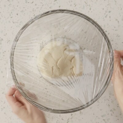Someone covering a glass bowl of dough with plastic wrap