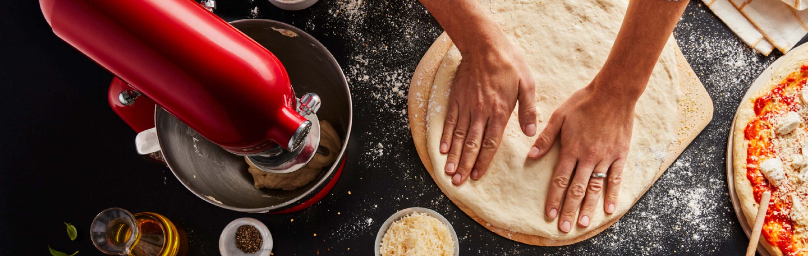Person flattening out pizza dough on countertop with KitchenAid® stand mixer and pizza ingredients
