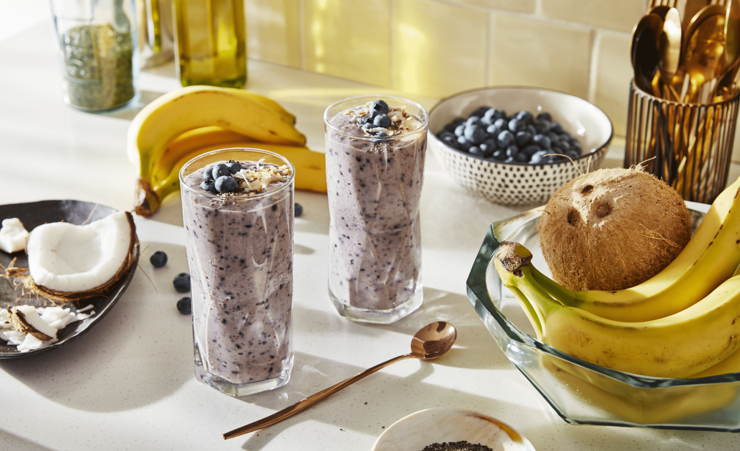 Two blueberry smoothies on countertop with bananas and coconut ingredients Two blueberry smoothies on countertop with bananas and coconut ingredients