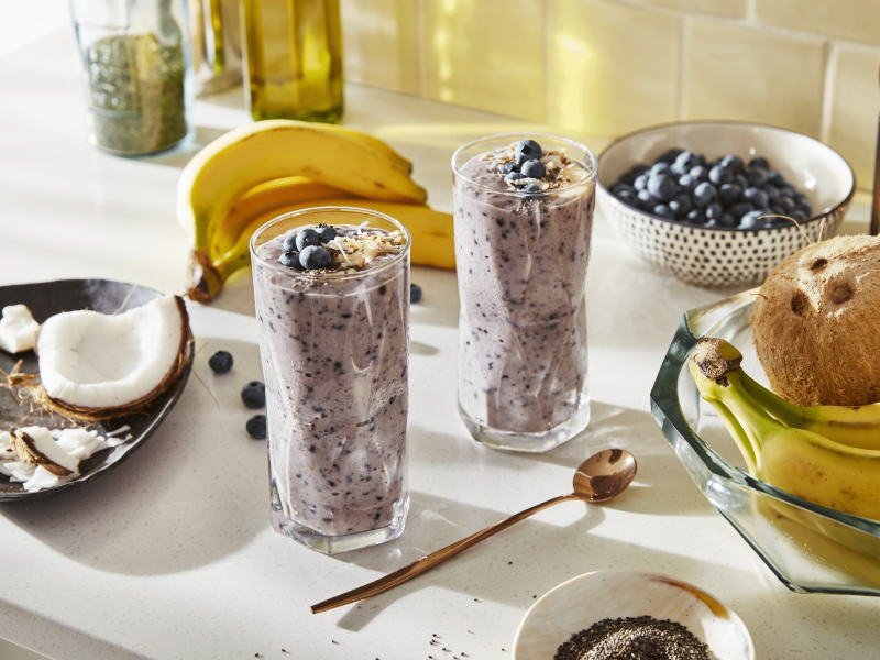 Two blueberry smoothies on countertop with bananas and coconut ingredients Two blueberry smoothies on countertop with bananas and coconut ingredients