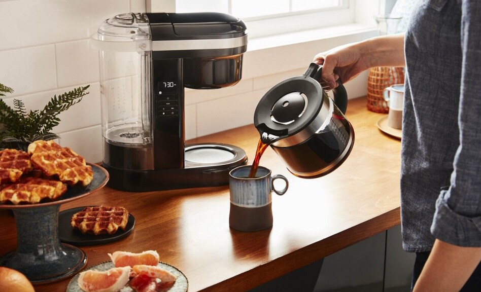 Person pouring coffee into a mug with coffee maker in background Person pouring coffee into a mug with coffee maker in background