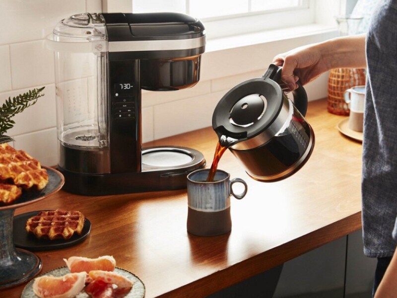 Person pouring coffee into a mug with coffee maker in background Person pouring coffee into a mug with coffee maker in background