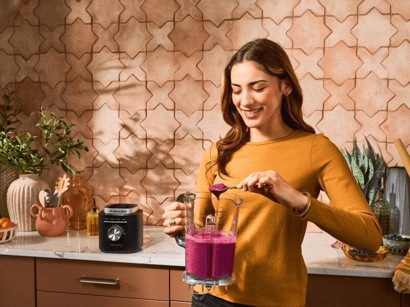 Woman with berry smoothie in a blender standing in front of wooden cabinetry and a white marble countertop
