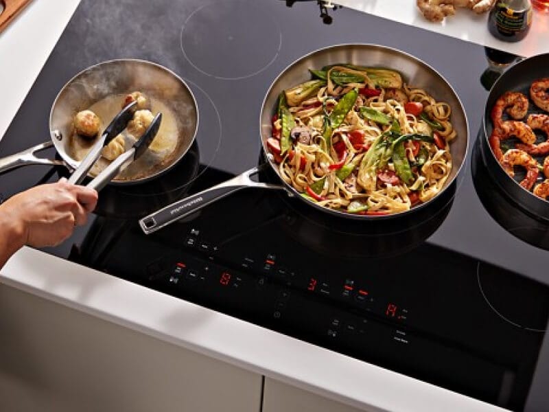 A person cooking pasta and shrimp on a black KitchenAid® electric induction cooktop A person cooking pasta and shrimp on a black KitchenAid® electric induction cooktop