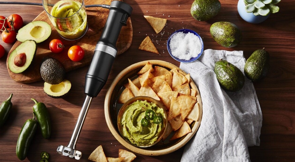 Immersion blender laying beside bowl of guacamole and chips surrounded by guacamole ingredients Immersion blender laying beside bowl of guacamole and chips surrounded by guacamole ingredients