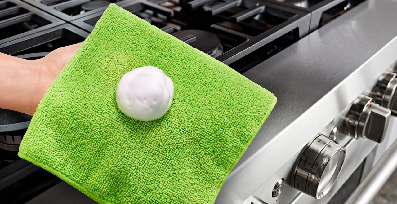 Close-up of a person's hand holding a green kitchen cloth with affresh® sprayed on it, over a commercial-style range. 