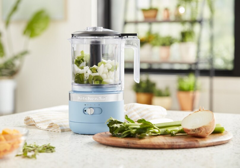 Blue food chopper with chopped onions and celery next to whole ingredients Blue food chopper with chopped onions and celery next to whole ingredients