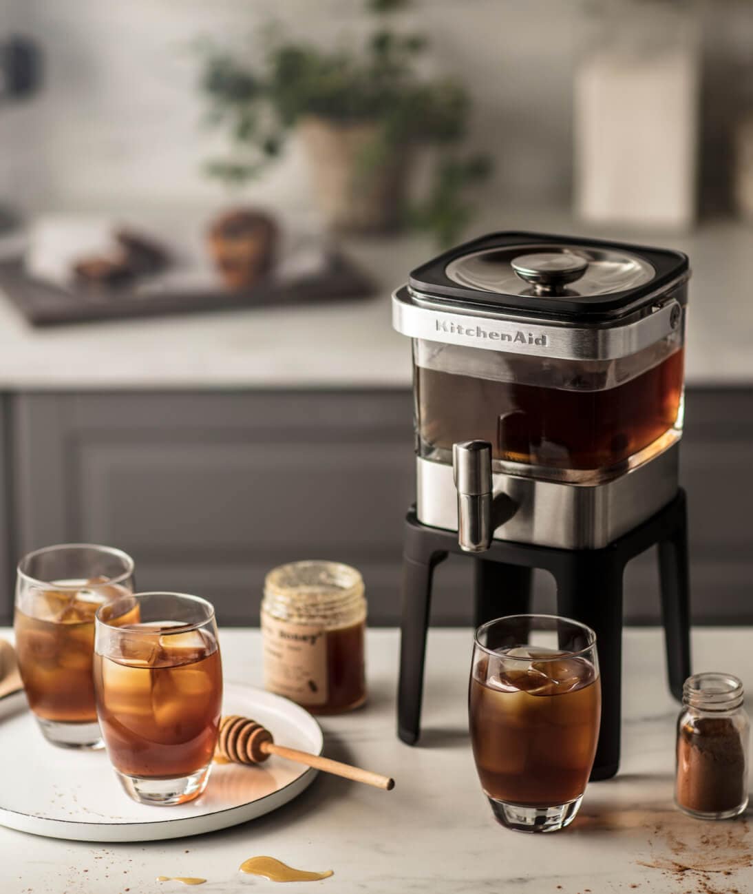 Cold brew coffee setup on kitchen counter including KitchenAid® cold brew coffee maker, three cups of cold brew, milk, honey and cinnamon.