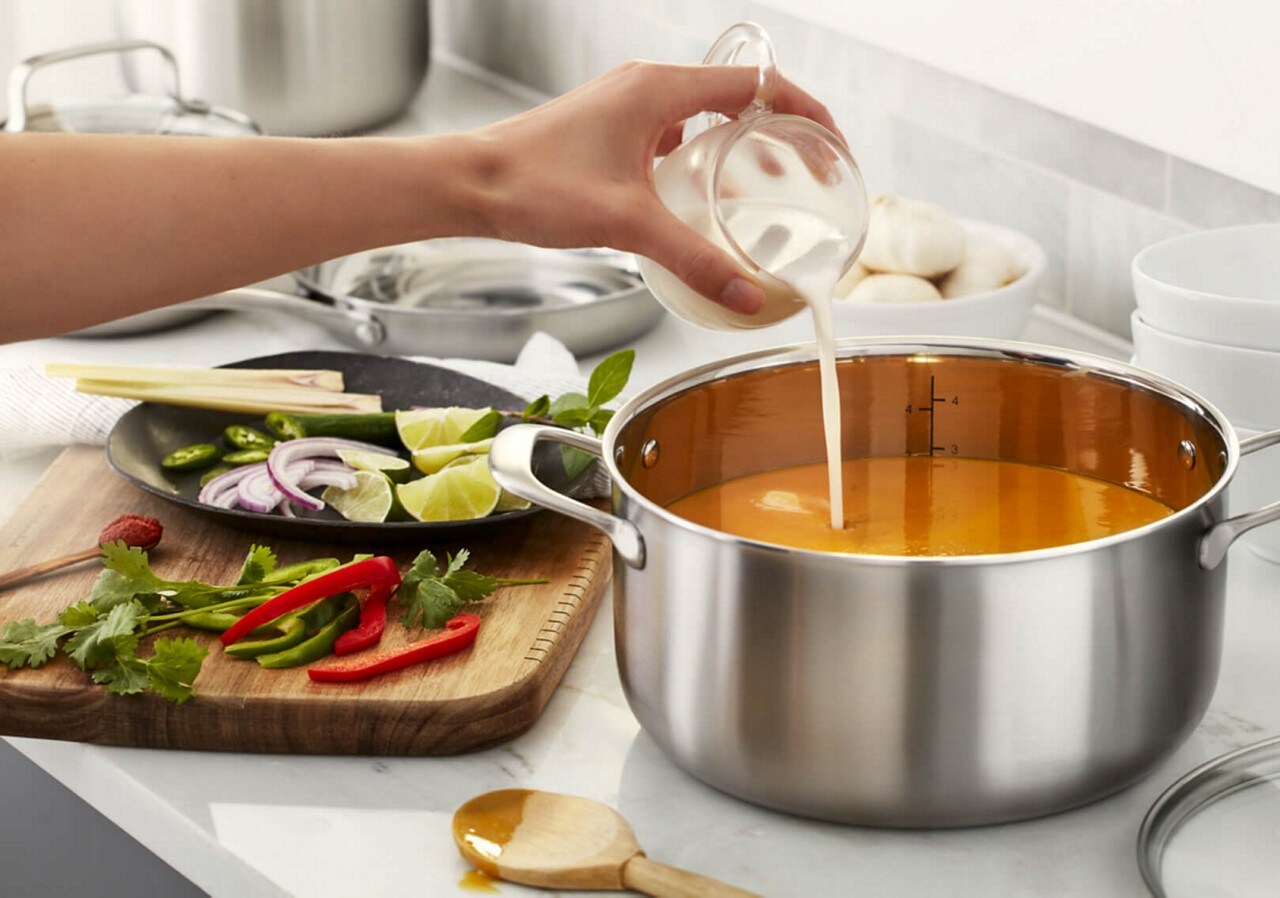Pouring cream into pan of soup sitting next to cutting board with herbs, peppers and lime wedges. Pouring cream into pan of soup sitting next to cutting board with herbs, peppers and lime wedges.